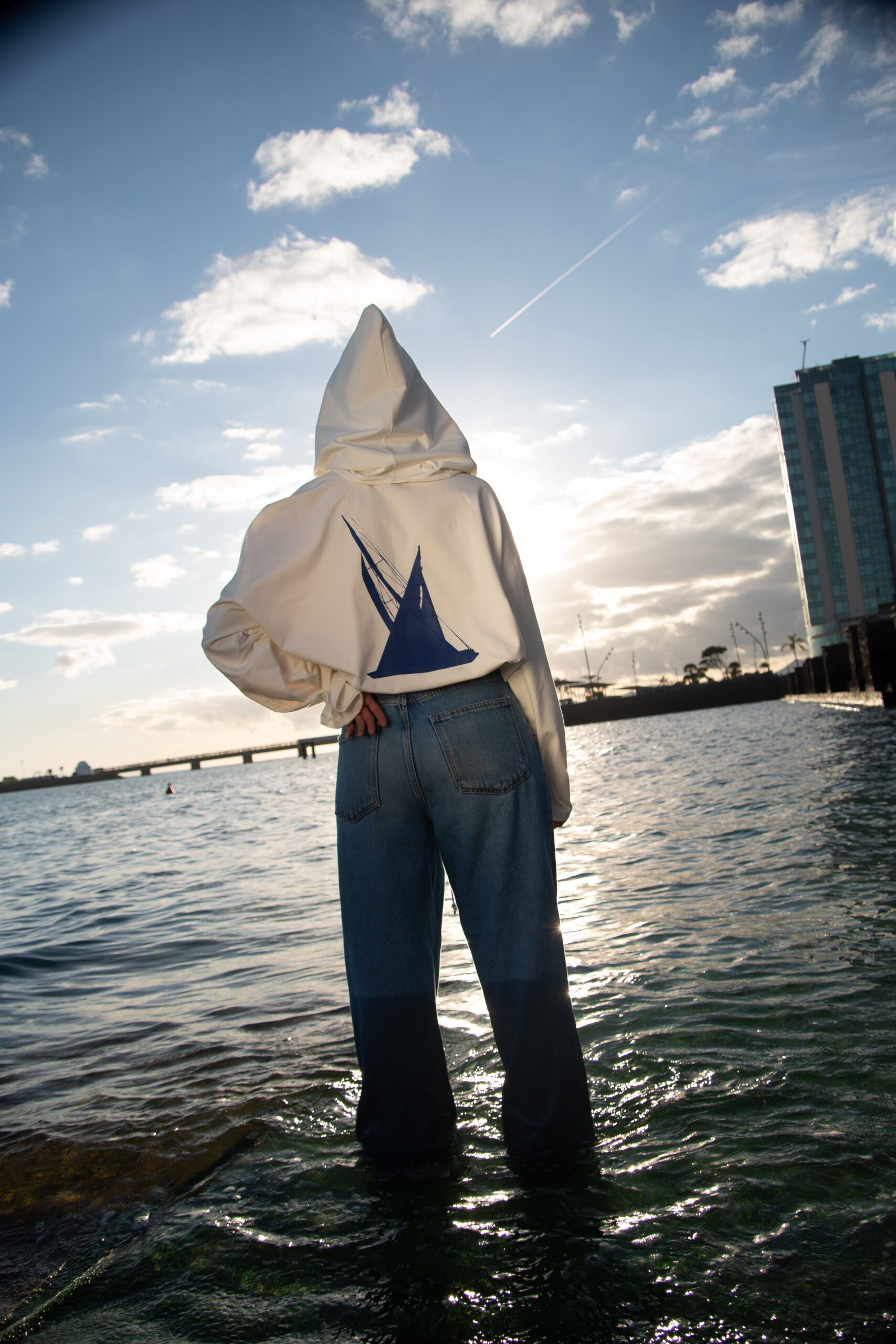 Persona de espaldas dentro del agua con la Spinnaker y la capucha puesta, a contraluz